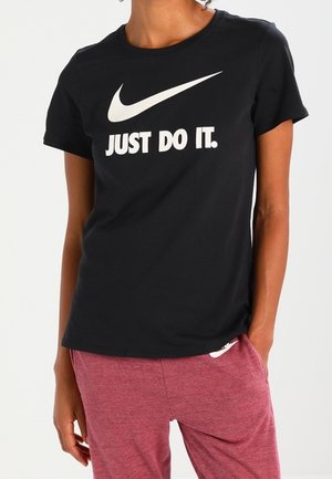Osoba ubrana w czarną koszulkę Nike z białym napisem i logo „Just Do It.”, w połączeniu z bordowymi spodniami, stojąca z jedną ręką w kieszeni.