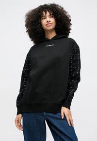 Γυναίκα με σγουρά μαλλιά που φοράει μαύρη μπλούζα με κουκούλα και μανίκια με υφή καρό, καθώς και μπλε τζιν, στέκεται μπροστά σε απλό ανοιχτό φόντο.