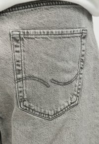 Κοντινό πλάνο μιας πίσω τσέπης από ανοιχτό γκρι τζιν με σκούρες ραφές σε ξεθωριασμένο τζιν, φορεμένο με λευκό πουκάμισο που είναι μπαλωμένο μέσα.