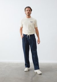Cream short-sleeve t-shirt with graphic on chest, paired with navy drawstring trousers. Model wears white sneakers on a gray floor.