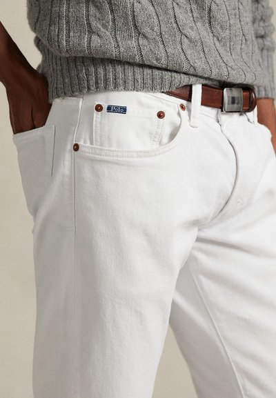 Calças de ganga brancas com corte justo, com uma etiqueta azul "Polo", detalhes castanhos e bolsos clássicos. Suéter cinza texturizado de malha de cabo visível.