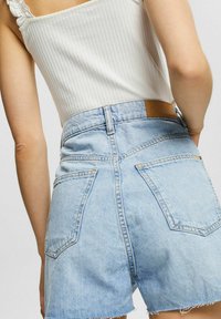 Pantalones cortos de mezclilla azul claro con un dobladillo deshilachado, que cuentan con bolsillos traseros y una etiqueta de cuero marrón en la cinturilla. Blusa blanca de canalé visible.