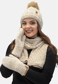 Gorro de punto beige con pompón, manoplas a juego y una bufanda texturizada. Todos los artículos presentan un patrón acanalado y un sutil detalle de marca.