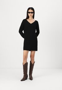 Abito maglione nero a coste con scollo a V, maniche lunghe e una silhouette aderente, abbinato a stivali da cowboy in pelle marrone con dettagli intricati.