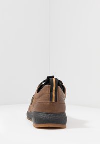 Baskets en cuir marron avec une semelle noire texturée, dotées d'une languette rayée et d'accents en mesh, vues de dos sur un fond uni.