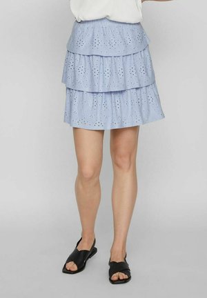 Kvinde iført en lyseblå, lagdelt nederdel med hulmønsterdetaljer og sorte flade sandaler med åbne tæer, stående mod en ensfarvet baggrund.