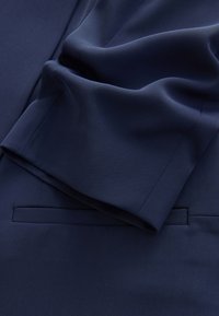 Tamnoplava tkanina presavijena, prikazuje krupni plan rukava jakne i džepa sa obrubom na odjeći.