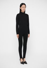 Pulover negru tricotat cu guler înalt, cu nervuri, asortat cu colanți negri mulați. Modelul poartă pantofi negri cu vârf ascuțit. Design minimalist cu siluetă elegantă.