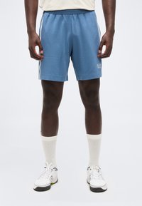 Άνδρας που φοράει μπλε αθλητικά σορτς με λευκές ρίγες στο πλάι, λευκές κάλτσες μέχρι τον αστράγαλο και λευκά αθλητικά παπούτσια, στέκεται μπροστά σε απλό φόντο.