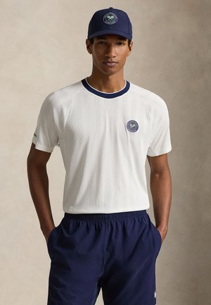 Młody mężczyzna w białej koszulce tenisowej z logo Wimbledonu, granatowych szortach i dopasowanej granatowej czapce, stojący z rękami w kieszeniach.
