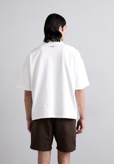 T-shirt en coton blanc à manches courtes, coupe décontractée, avec un petit détail en texte noir dans le dos. Porté avec un short marron.