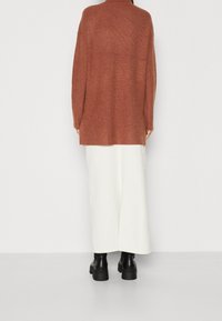 Cardigan lungo a maglia color ruggine con una texture a coste, dotato di un collo a camicia e spalle calate, indossato sopra una maxi gonna bianca.