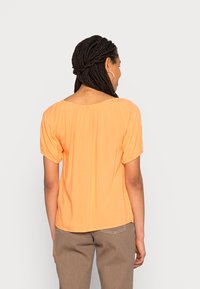 Orange top med en afslappet pasform, korte puffærmer og en let rynket halsudskæring. Glat stof med en subtil glans, parret med brune bukser.