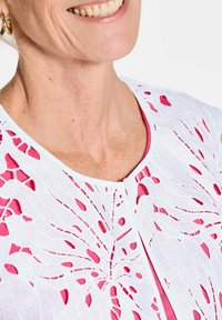 Weißes Kleidungsstück mit filigranem, durchbrochenem Blumenmuster und rosa Akzenten, runder Halslinie und Knopfverschluss entlang der Knopfleiste.