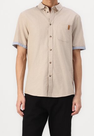 Beżowa koszula na krótki rękaw z guzikami, kieszonka na piersi, jasnoniebieskie mankiety, noszona z czarnymi spodniami.