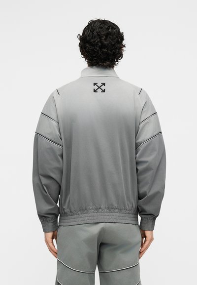 Άνδρας με σγουρά μαλλιά, φοράει γκρι μπουφάν και παντελόνι με ραμμένες οριζόντιες γραμμές, γυρισμένος προς τα πίσω σε απλό φόντο.