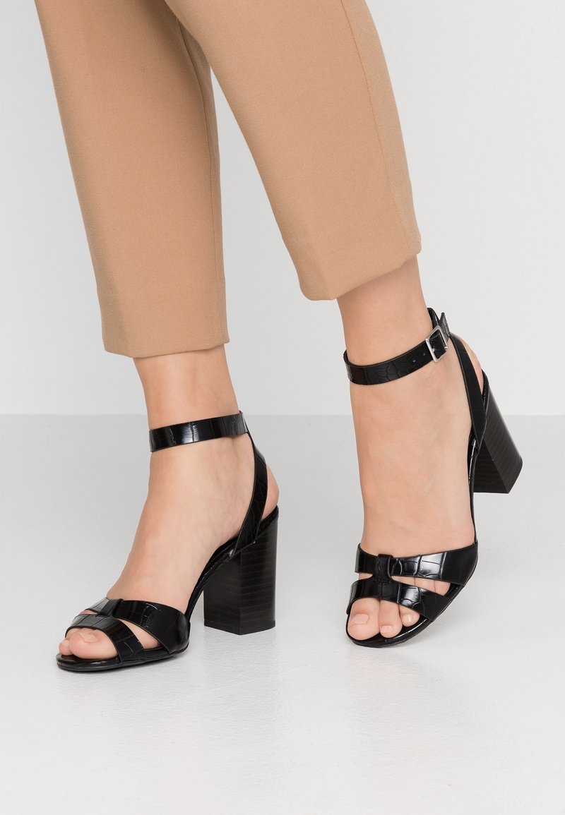 Pieds chaussés de sandales à talons hauts noires avec brides et boucles à la cheville, associées à un pantalon beige raccourci, debout sur un sol blanc.