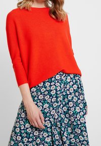 Jasnopomarańczowy sweter z dzianiny o luźnym kroju i rękawach do trzech czwarte, w połączeniu z granatową spódnicą we florystyczny wzór z białymi stokrotkami i zielonymi liśćmi.