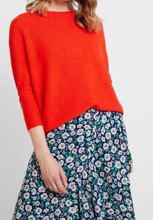 Chandail tricoté orange vif à coupe ample et manches trois quarts, associé à une jupe florale bleu marine ornée de marguerites blanches et de feuilles vertes.