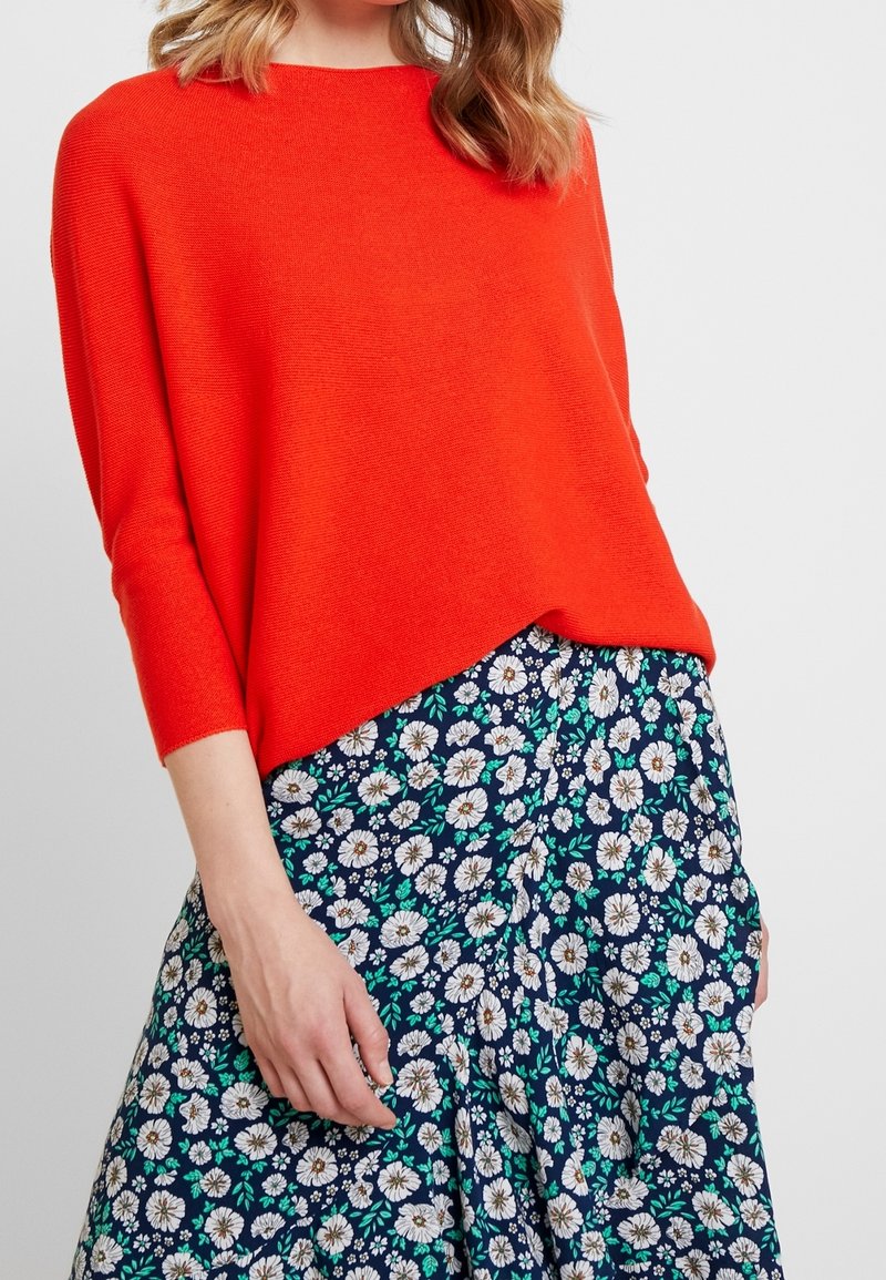 Jasnopomarańczowy sweter z dzianiny o luźnym kroju i rękawach do trzech czwarte, w połączeniu z granatową spódnicą we florystyczny wzór z białymi stokrotkami i zielonymi liśćmi.