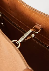 Interno di una borsa in pelle color tan con zip metallica argentata e chiusura a clip. Presenta superfici testurizzate e dettagli hardware brandizzati.