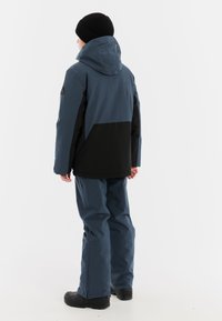 Blauwe en zwarte waterdichte jas met capuchon, voorzien van een contrasterend kleurvlakdesign, een soepele textuur en zwarte broek.