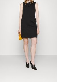 Černé bezramenné šaty s drapeovaným výstřihem a rovnými liniemi. Doladěné se žlutou kabelkou a černými sandály na vysokém podpatku. Hladký textilní povrch.