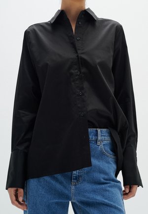 Personne portant une chemise noire asymétrique à boutons avec de larges poignets associée à un jean bleu, cadrée à mi-torse sur fond uni.