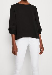 Haut noir texturé à manches trois-quarts, avec des détails froncés et des accents en dentelle. Associé à un pantalon blanc, la tenue présente une coupe décontractée.