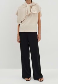 Beige texturerad kortärmad topp med en knytning vid halsen, kombinerad med svarta vida byxor och platta sandaler.