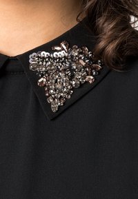 Col haut de vêtement noir avec des embellissements complexes, comprenant des perles et des sequins de différentes tailles et couleurs, créant un effet décoratif texturé.