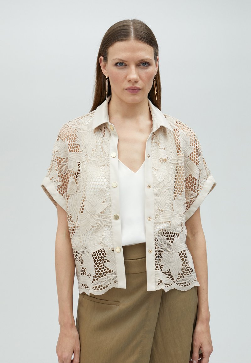 Beige shirt with lace overlay, short sleeves, button-down front, and a collar. Worn over a white top and paired with khaki pants.