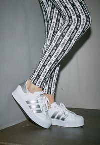 Bílé kožené tenisky se stříbrnými akcenty, s texturovanou špičkou a třemi stříbrnými pruhy. V kombinaci s legínami s černobílým potiskem loga.