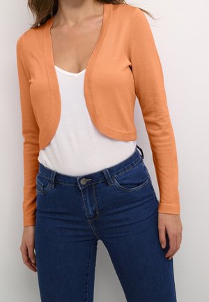 Femme portant un cardigan court orange ouvert devant, sur un t-shirt blanc à col en V et un jean bleu taille haute, debout devant un fond uni.