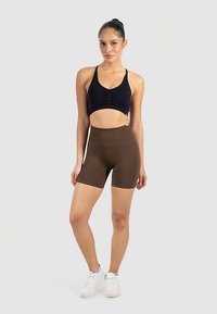 Svart sportig croptopp med rynkad detalj, kombinerad med höga bruna shorts, bärs med vita sportskor mot en neutral bakgrund.