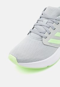 Chaussure de sport grise avec une tige en maille texturée, des accents vert clair et une semelle blanche. Présente des lacets plats et un design épuré.