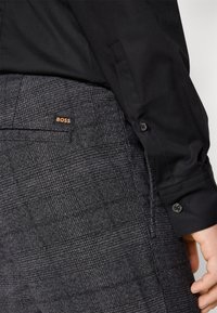 Gros plan d'une personne portant un pantalon gris foncé à carreaux avec une étiquette logo BOSS et une chemise noire à manches longues avec des boutons sur le poignet.