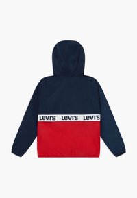 Huvjacka i marinblått och rött. Har elastiska ärmslut, ett vitt märkesband med "Levi's" och ett mjukt, lätt material.