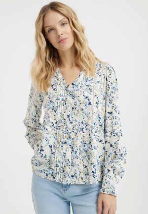 Blusa floreale con scollatura a V, fondo bianco e motivi blu, gialli e verdi. Pieghe dettagliate e polsini elastici aggiungono texture.
