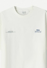 Bílý bavlněný tričko s krátkým rukávem. Obsahuje modrý text, včetně "RIO Ipanema Soul" a "saudade for Brazil." Jednoduchý design, hladká textura.