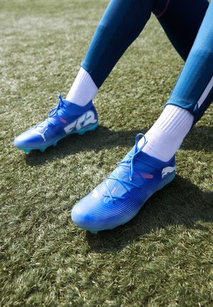 Blauwe voetbalschoenen met een gemêleerd mesh bovenwerk, voorzien van veters en een wit logo. De zool heeft een turquoise kleur en gemolde noppen voor grip.