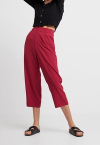 Cranberrykleurige cropped broek met hoge taille, voorplooien en een gladde afwerking. Gecombineerd met een zwart geribbeld knoopshirt en zwarte sandaaltjes.