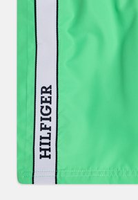 Gröna badshorts i lätt material med ett vitt rand och texten "HILFIGER" i svart, nära sidans söm.