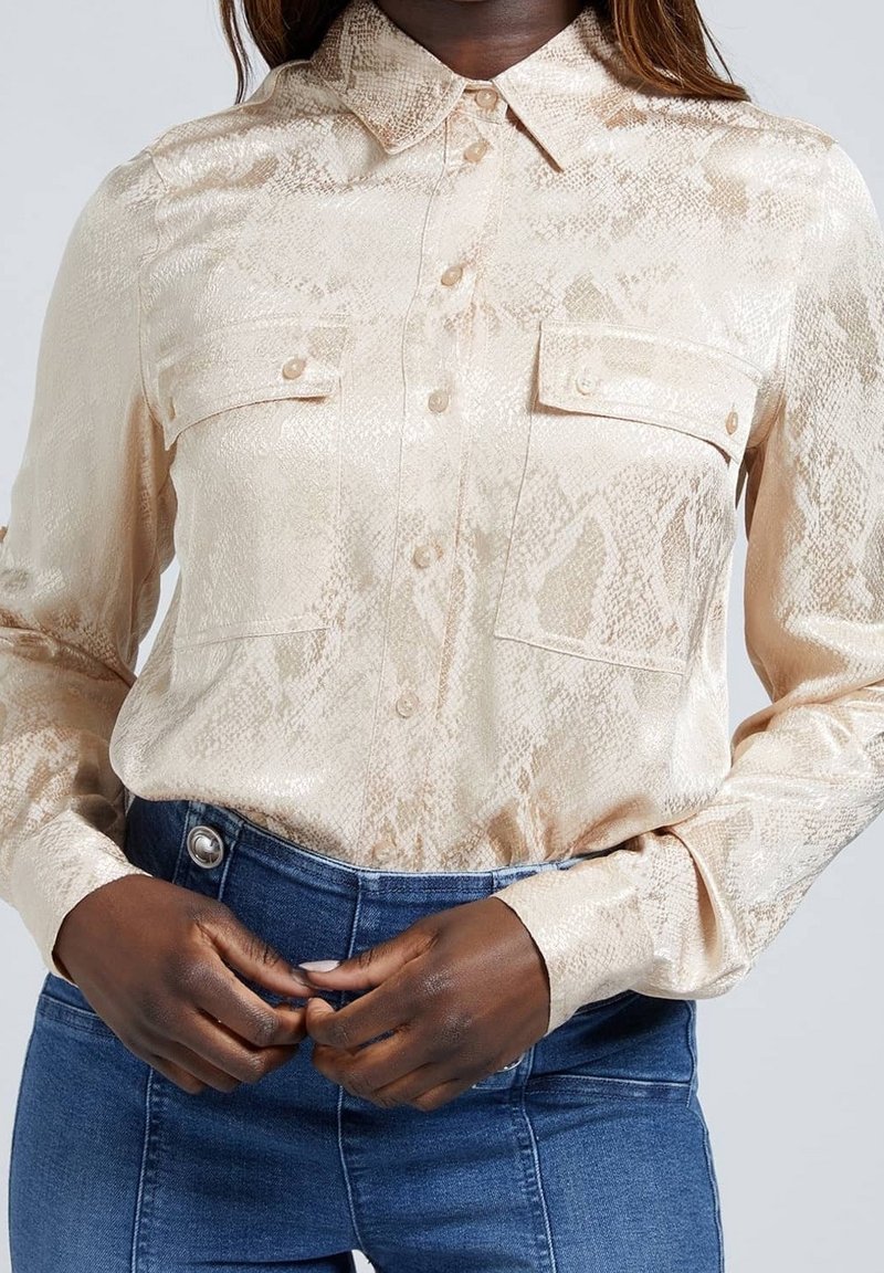 Femme portant une chemise beige imprimé serpent rentrée dans un jean bleu taille haute, mains ajustant la taille.