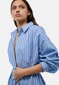Chemise bleu clair à boutons, avec des rayures verticales, dotée d'une poche poitrine et de manches relaxées resserrées aux poignets. Texture de tissu douce.