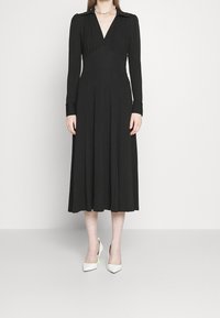 Robe noire à manches longues avec un décolleté en V et un col, jupe évasée et taille plissée. Associée à des escarpins blancs à bout pointu avec un accent néon.