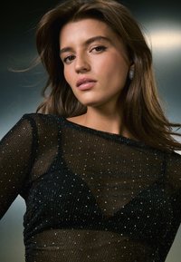 Melnā caurspīdīgā blūze ar teksturētu rakstu un smalkiem mirdzumiem, kas papildina kārtaino dizainu. Modelim ir garas brūnas matu un auskari ar studiem.