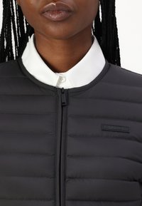 Črn napihnjeni jopič z vodoravnimi gebrati, z zadrgo in belo srajco pod njim. Logotip Calvin Klein na prsih.