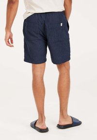 Marineblauwe shorts met een gestructureerde afwerking, voorzien van een elastische tailleband en twee achterzakken, gecombineerd met donkere slippers.