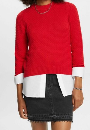 Femme portant un pull rouge texturé par-dessus une chemise blanche aux poignets retroussés, associée à une jupe en denim noire à ourlet effiloché et un bracelet en argent.